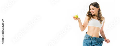 Fotomural smiling slim young woman in big jeans holding ripe green apple isolated on white