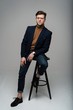 © opolja - full length portrait of a young fashion man sitting on a chair and looking into the camera on a gray background.