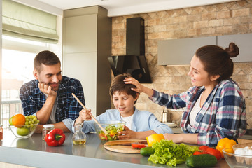 Wall Mural - Family at home leaning on table in kitchen together parents looking at son mixing salad smiling proud