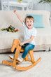 © LIGHTFIELD STUDIOS - adorable excited preschooler boy riding rocking horse and gesturing with hand at home