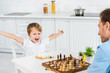 © LIGHTFIELD STUDIOS - preschooler son cheering with hands in air while playing chess with father at home