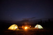 © anatoliy_gleb - Camping in mountains in the evening. Bright bonfire burning between two tourists, man and woman sitting opposite each other near illuminated tents under night starry sky on distant hills background.