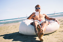 Handsome Man In Sunglasses And Swimming Pants Resting On Pouf On Beach With Cup Of Tea Or Coffee, In The Background Windsurfers In The Sea. Active Lifestyle. Focus On Feets