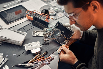 Wall Mural - Serviceman uses magnifier and screwdriver to repair damaged smartphone in the electronic workshop.