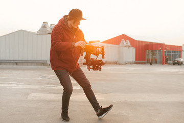 Man with a stabilizer in his hands makes a video on a mirrorless camera. Portrait of the video operator in full height, takes video on the background of the sunset. Process of creating a video