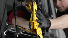 Professional Worker Guy (Man) Wipes The Car After Painting (ceramics) In A Special Uniform With Black Gloves And With A Yellow Rag. Concept Of: Young Master, Preparing For The Sale, Cleaning.