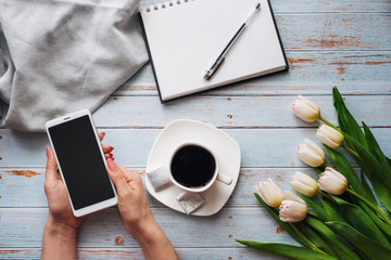 Wall Mural - bouquet of white tulips with a Cup of coffee and a smartphone in women's hand and an empty notebook on a wooden table