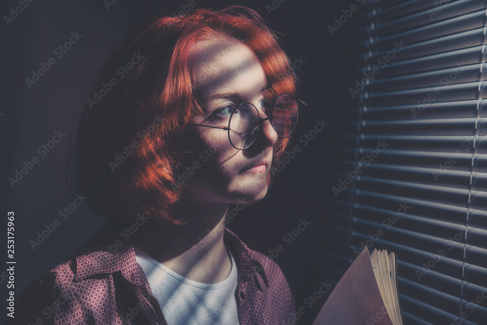 Red-haired girl with glasses in a plaid shirt and a white t-shirt is sad at the window with blinds - obrazy, fototapety, plakaty 