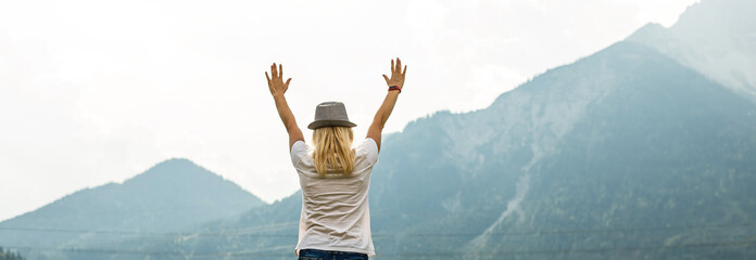 Young woman with backpack standing on cliff's edge and looking to a sky with raised hands