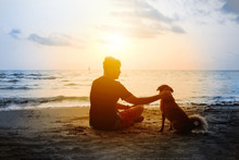 Man And Dog Sitting On Sea Beach And Looking At Sunset. Friendship And Dedication Between Human And Home Pet Concept. Outdoor Walking With A Dog.