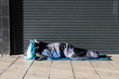 Homeless person asleep in a sleeping bag on a pavement sidewalk in front of a metal shutter.  Face not visable.