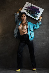 Youns model artist wearing jean jacket,black trousers and yellow high heel shoes.standing on background and posing with her painting art canvas