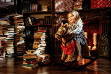 Beautiful Little Girl In A Blue Dress Like A Princess Is Sitting On A Toy Wooden Horse In A Vintage Studio.