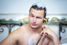 Close Up Portrait Of Funny Sexy Man Having Fun Relaxing In Shower Taking Bath With Foam, Fooling Around, Talking On The Phone With A Serious-faced Vintage Shower Head