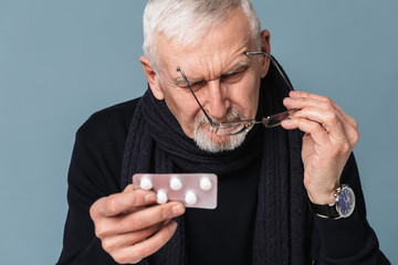 Old pensive sick man with gray hair and beard in eyeglasses and scarf thoughtfully looking on pills over blue background isolated