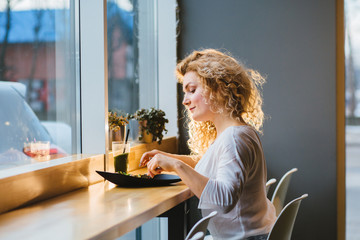 Wall Mural - Beautiful curly blond american woman eating salad in a restaurant and looking in the window. Focus on a salad plate. Lunch or dinner with a healthy meal at the restaurant