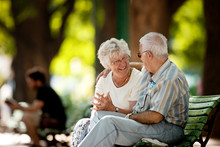 Senior Woman Smiles At A Senior Man With His Arm Around Her Shoulders As They Sit Together On A Park Bench.