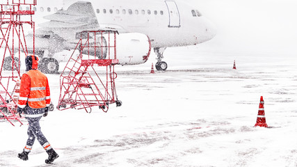 Wall Mural - heavy snow storm at winter airport and maintenance person walking on front of passenger airplane on white snow covered runway bad weather snowfall on airfield flight delay cancelled air travel scene