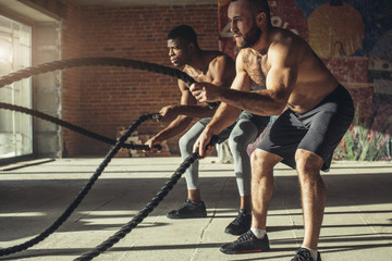 Wall Mural - Muscular half-naked body athletes doing some crossfit exercises with a rope indoor, preparing to competitions and effectively burning excess fat.