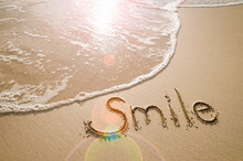 Smile Message Handwritten On The Smooth Sand Of An Empty Beach With An Oncoming Wave And Sunlight Lens Flare