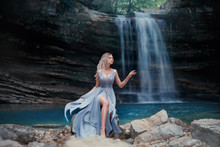 A Curly Blonde Girl In A Luxurious Blue Dress Sits On White Stones Against The Backdrop Of A Fabulous Landscape. River Mermaid Near The Lake With A Waterfall. Art Photography