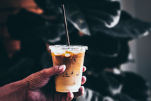 Man Holding Plastic Glass Of Iced Coffee With Milk On Blurred Background With Cream Poured Over And Coffee Beans On A Nature Green Leave Dark Tone. Cold Summer Drink Background