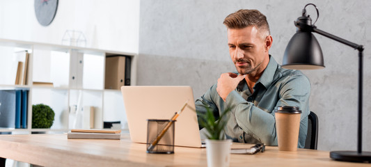 panoramic shot of pensive businessman sitting near laptop in modern office