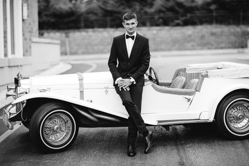 Wall Mural - Elegant fiance in a black suit. Man standing near old car