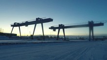A Large Gantry Cranes Working On The Rail While Automobile Cranes Worki Behind It, Time Lapse. Gantry Crane In The Industrial Zone In Winter Morning On Rising Sun Background.