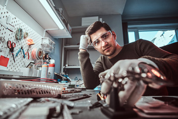 Wall Mural - Stylish electronic technician in goggles, leaning on a desk in a repair shop, looking at a camera with a thoughtful look