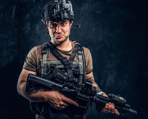 Man in t-shirt with tattoo on his hand wearing body armor and helmet with a night vision holding assault rifle and looking at a camera with a funny look. Studio photo against a dark textured wall