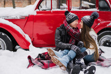 Cheerful Couple In Warm Cozy Clothes Near Red Vintage Car Covered With Snow In The Garden. Cold Happy Winter Day. Holidays, Christmas, New Year, Winter, Love, Beauty Concept.
