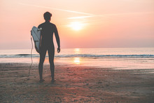 Male Surfer Standing On The Beach Waiting For Waves At Sunset Time - Man With Surfboard Wearing Wet Suit Looking The Waves - Extreme Sport Concept