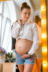 Pregnant happy woman touching her belly. Pregnant young mother portrait, caressing her belly and smiling. Healthy Pregnancy concept, brunette expectant female near the window