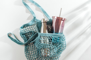 Mesh bag with reusable metal water bottle and cup on white background. Sustainable lifestyle. Zero waste concept. No plastic.