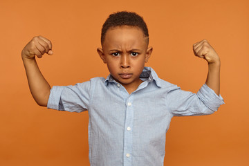 Wall Mural - Serious seven year old dark skinned boy having grumpy facial expression, frowning and tensing biceps, demonstrating his strong arms, being full of energy. Strength, vitality, health and well being