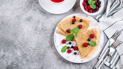 Delicious Crepes Breakfast on gray concrete table background. Orthodox holiday Maslenitsa. Pancakes with berry black currant, raspberry, jar of honey and mint. White cup of tea. Banner