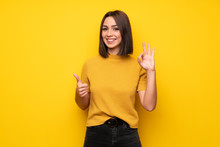 Young Woman Over Yellow Wall Showing Ok Sign With And Giving A Thumb Up Gesture