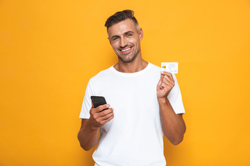 Wall Mural - Image of cheerful guy 30s in white t-shirt holding mobile phone and credit card