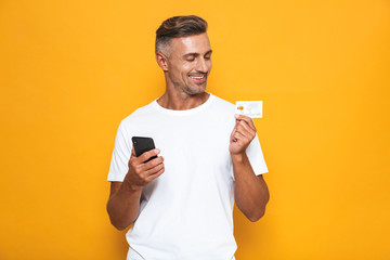 Wall Mural - Image of positive guy 30s in white t-shirt holding mobile phone and credit card