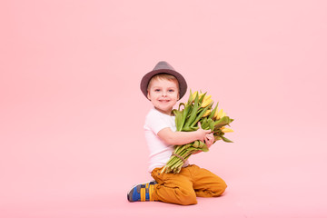 Wall Mural - Adorable smiling child with spring flower bouquet looking at camera isolated on pink. Little toddler boy in hat holding yellow tulips as gift for mom. Copy space for text 