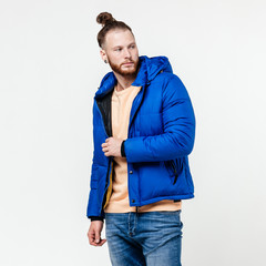 Wall Mural - Fashionable man with beard and bun hairstyle dressed in yellow sweater, jeans and blue down jacket poses in the studio on the white background