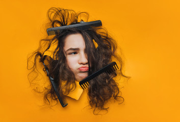 A funny teenage caucasian girl with problematic curly hair expresses displeasure on her face. Three black combs tangled in hard hair on yellow paper background.