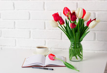 Tulip Flowers Are In A Vase On The Table, Cup Of Coffee And Diary, White Brick Wall As Background