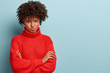 Offended black woman purses lips, keeps hands crossed, feels upset and dissatisfied, wears red casual jumper, looks with disappointment, stands over blue wall with empty space for your text.