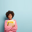 © wayhome.studio  - Photo of hesitant thoughtful Afro American student prepares for making home assignment, focused upwards, holds papers and textbooks, purses lips, wears transparent spectacles and oversized jumper