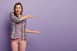 © WHstudio Leushin N - Studio shot of happy shocked young woman shapes big object with both hands, expains what pile of papers she has to look through, poses over purple wall, wears checkered shirt, rosy trousers.