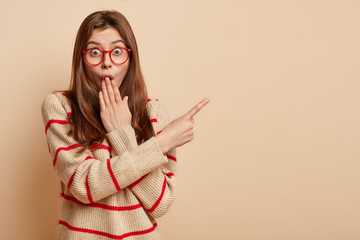 Wall Mural - Shocked young woman covers mouth not speak, points away with fore finger, shows something amazed, wears red rim glasses and casual sweater, stands over brown wall with mock up space for your text