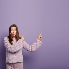 Sticker - Photo of surprised European woman has shocked expression points aside with both fore fingers, opens mouth from wonder, demonstrates something amazing, wears knitted jumper, isolated on purple