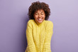 © wayhome.studio - Indoor shot of cheerful lady with curly hair, eyes shut, has toothy smile, shows perfect teeth, listens confession in love from boyfriend, wears yellow sweater, isolated over purple background